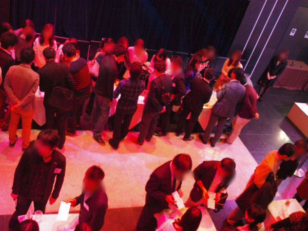 赤い照明が特徴的な屋内のイベント会場で、多くの人々が立ち話をしている様子が写っています。参加者の中にはスーツ姿のビジネスパーソンもおり、交流会やパーティーのような雰囲気です。