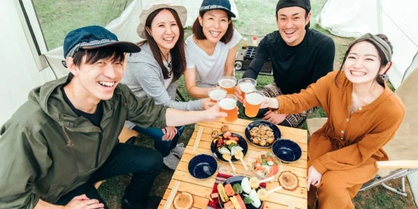 屋外で友人グループがテーブルを囲み、ビールを片手に乾杯している楽しそうな様子です。串焼きやサラダなどの料理も並び、笑顔で会話を楽しんでいます。
