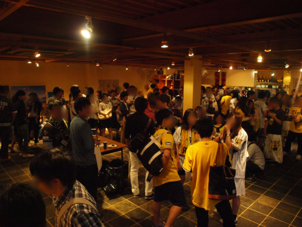 阪神タイガースのファンらしき人々が屋内のイベント会場に集まり、交流を楽しんでいる様子。多くの参加者がチームカラーの黄色や縦縞の服を着用している。暖色系の照明が灯るカジュアルな雰囲気の社交イベントである。