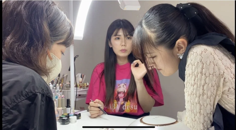 3人の女性がネイルをしている様子を捉えた画像です。中央の女性がネイルブラシを持ち、手元の作業に集中しており、右側の女性はそれを熱心に覗き込んでいます。左側の女性はマスクを着用し、同様に手元を見ています。ネイル用品やUVライトがテーブルに置かれています。