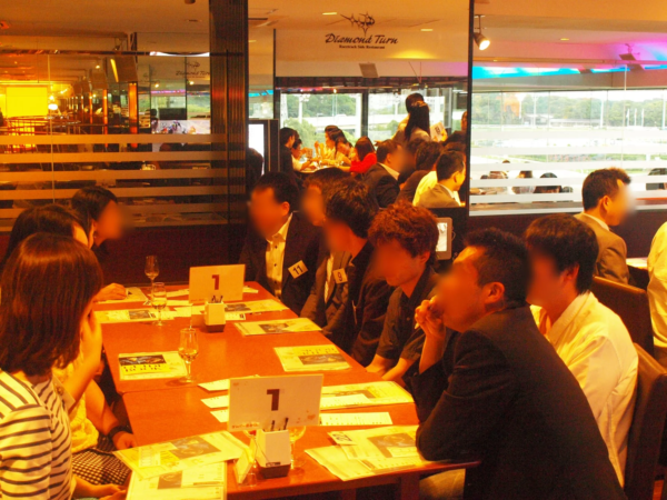 競馬場に隣接するレストラン「Diamond Turn」で、多くの人々がテーブルを囲んで会食やイベントに参加している様子。窓からは競馬場のコースが見えます。