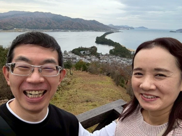 笑顔の男女が、日本三景の一つである天橋立を背景に自撮りしている様子。