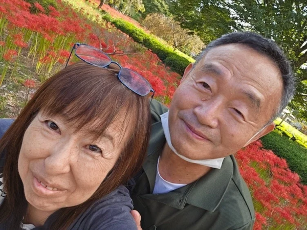 赤い彼岸花が咲き誇る中で、笑顔の夫婦が自撮りをしている。