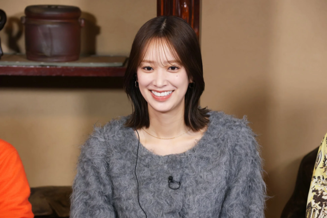 ふわふわした灰色のセーターを着た若い女性が、明るい笑顔でカメラを見つめています。首元には華奢なネックレスとラベリアマイクが見え、温かみのある室内で撮影されたポートレートです。