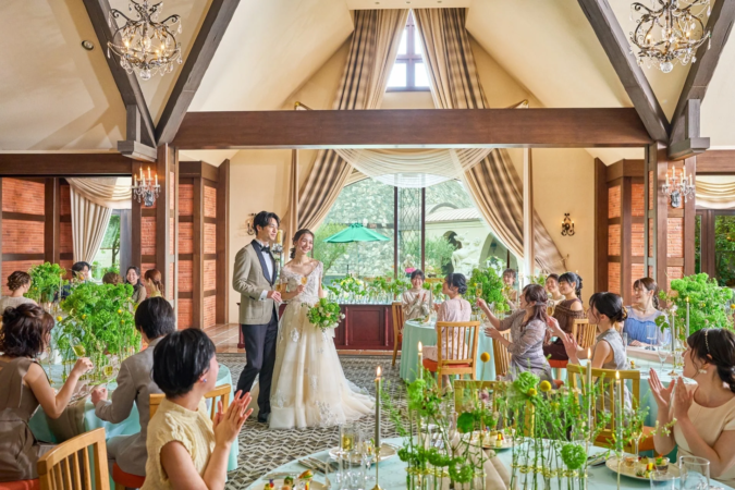 結婚式の披露宴で、新郎新婦がゲストに囲まれ、拍手と笑顔で祝福されている様子です。会場は緑豊かな装飾とシャンデリアで飾られ、大きな窓からは自然光が差し込む明るい雰囲気です。