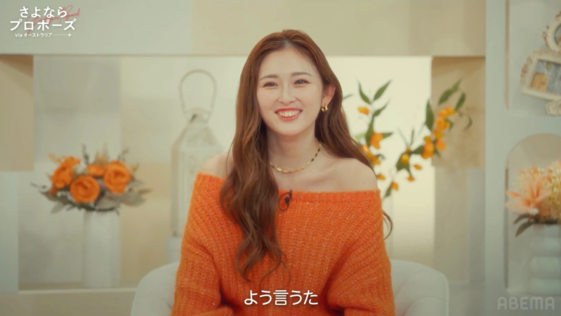 オレンジ色のオフショルダーセーターを着た笑顔の女性がカメラを見ています