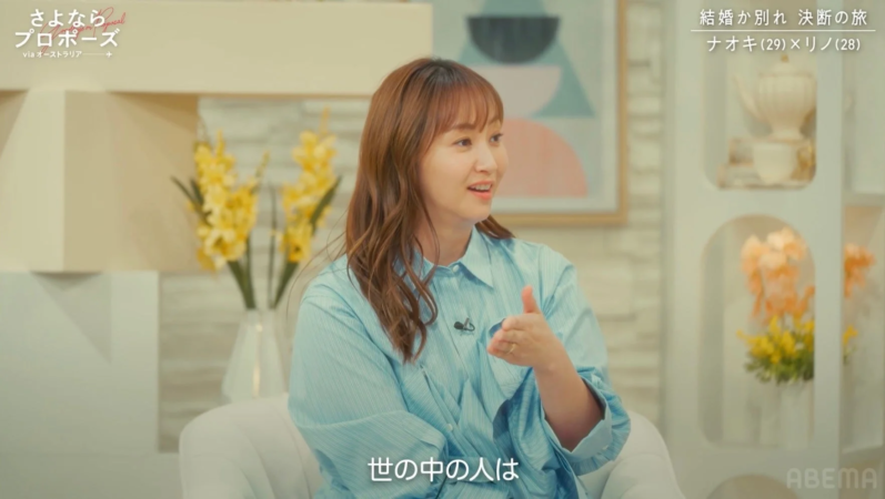 テレビ番組のスタジオで、茶色の髪の女性が水色のシャツを着て話している様子