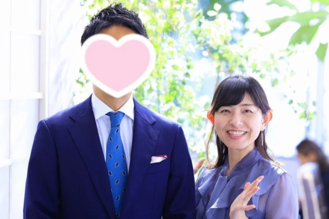 青いスーツ姿の男性(顔はハートで隠されている)と、笑顔でカメラを見つめる紫色のブラウスを着た女性のツーショットです。