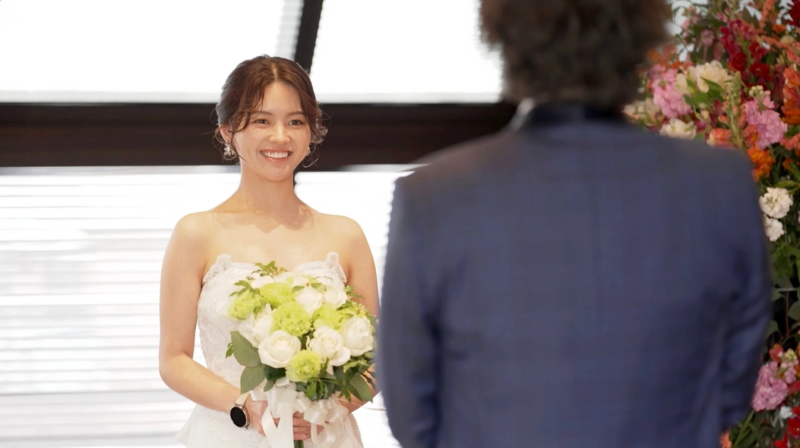結婚式の最中、純白のウェディングドレスを着た花嫁が、白いバラと緑のブーケを持ち、幸せそうな笑顔で新郎を見つめている瞬間を捉えた画像です。背景には色とりどりの花が飾られています。