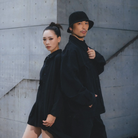 黒いシャツとパンツを着用した男女が、コンクリートの壁を背景に背中合わせに立つポートレート