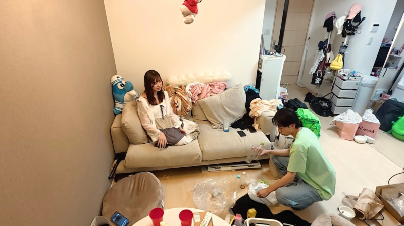 散らかったリビングで女性がソファに座り、男性が片付けをする様子