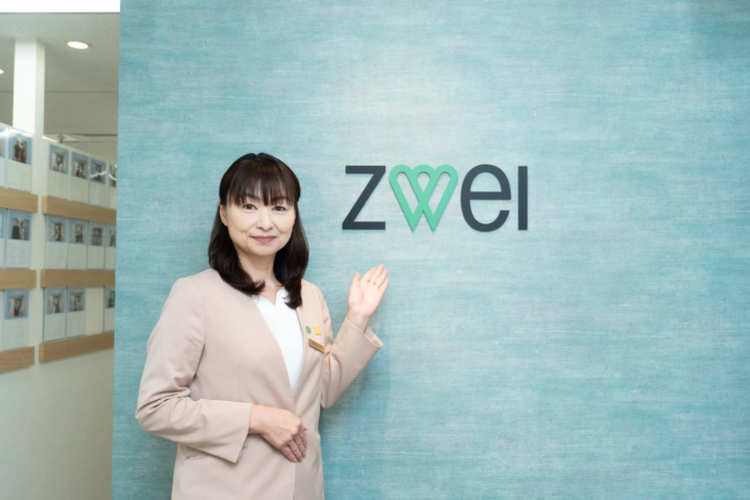 青い壁の前に立つ笑顔の女性が、結婚相談所「zwei」のロゴを指差して案内しています。オフィスのような空間で、親しみやすい雰囲気です。