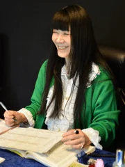 緑色のカーディガンを着た長い黒髪の女性が、書類を前にして笑顔を見せています。
