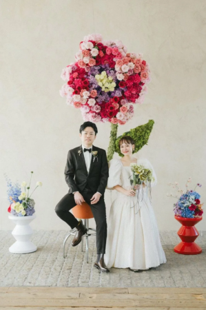 カラフルな巨大な花のアートを背景に、タキシード姿の新郎とウェディングドレス姿の新婦が並んでポーズをとっています。