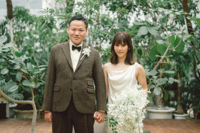 緑豊かな屋内庭園のような場所で、新郎新婦と思われるカップルが手をつないでカメラを見つめています。