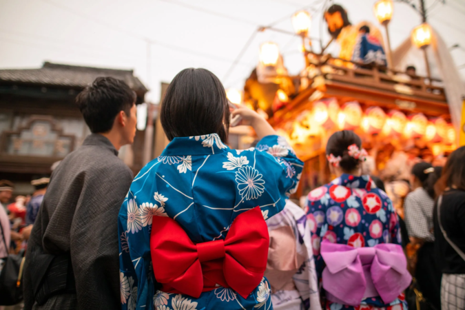 日本の伝統的な祭りの風景。浴衣や着物を着た人々が、提灯で飾られた豪華な山車を見上げている後ろ姿が捉えられています。