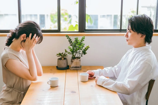 夫婦がテーブルを挟んで座っており、女性は顔を覆って悲しげな様子。